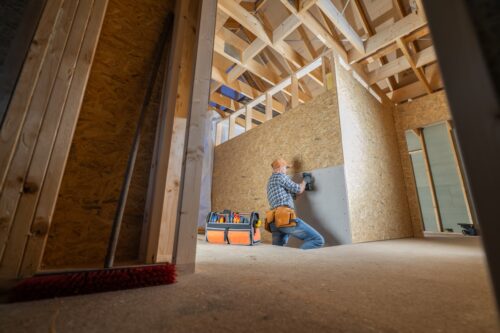 Inside a residential building under construction, a worker is focused on installing drywall on a newly framed wall. With tools organized nearby, the worker is using a power tool to secure the drywall into place, contributing to the progress of the ongoing project.